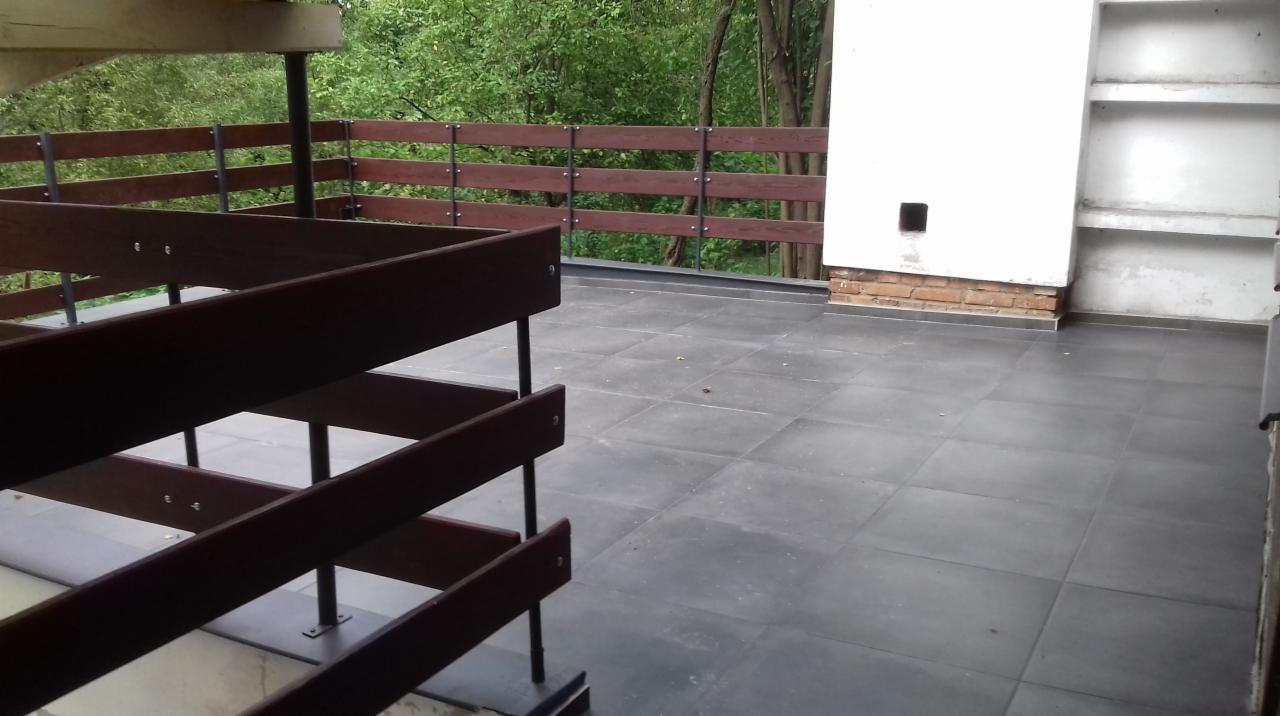 Wykończony taras z szarymi płytkami, drewnianą balustradą z metalowymi elementami i fragmentem białej ściany z ceglanym wykończeniem u dołu.