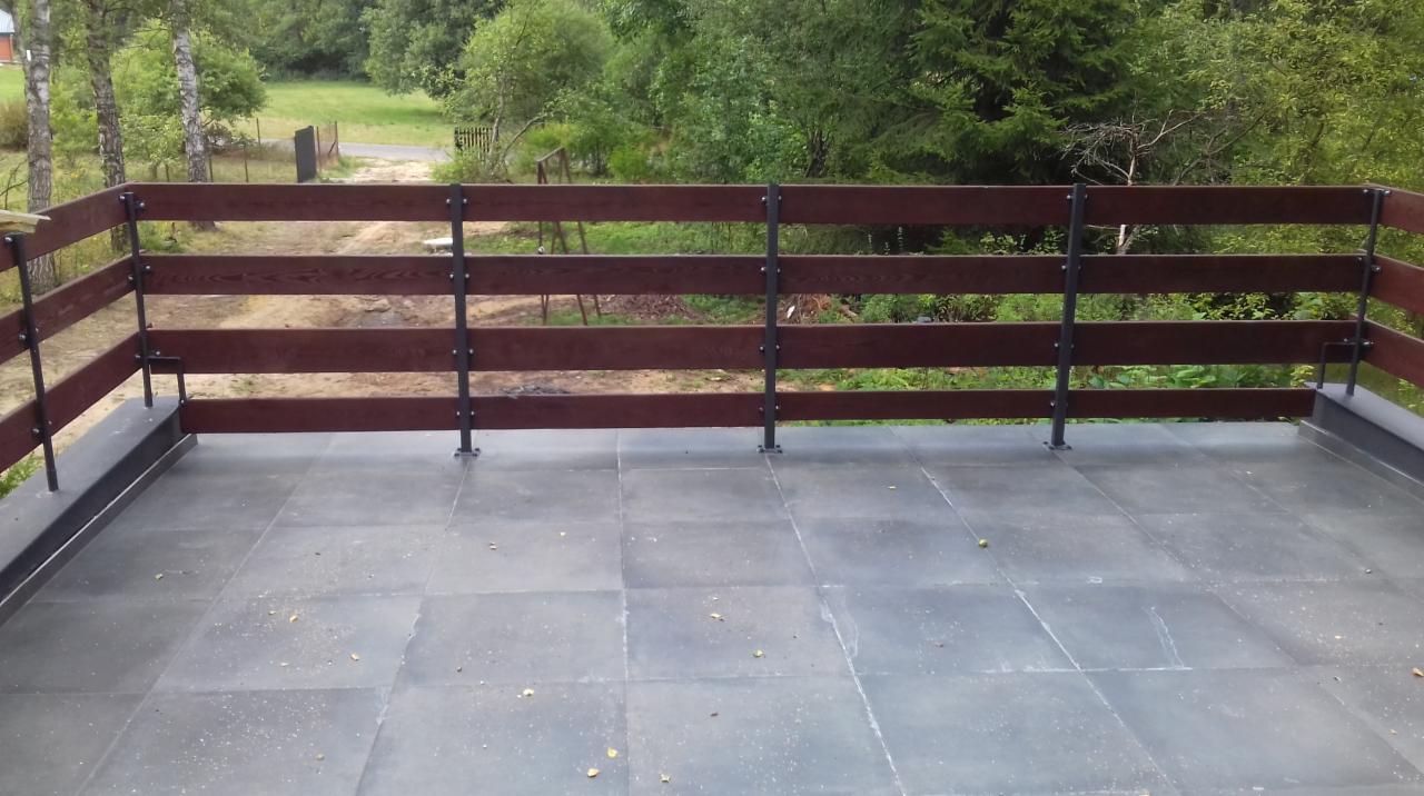 Zadaszony taras z drewnianą balustradą i szarą podłogą z płytek na tle zieleni drzew.