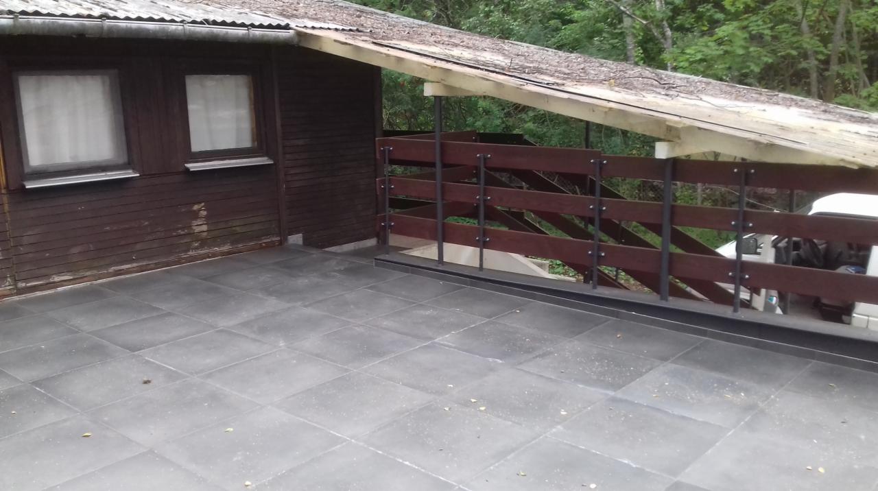 Zewnętrzny taras wyłożony szarymi płytkami, z drewnianą balustradą z metalowymi słupkami, przylegający do brązowego budynku z dwoma oknami i zadaszeniem z blachy falistej.