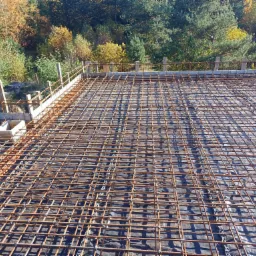 Zbrojenie stropu ułożone na czarnej folii, widoczne drewniane szalunki i otaczający krajobraz leśny w jesiennej scenerii.