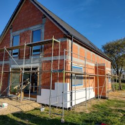 BARTZ Bartłomiej Wylężek - Surowy budynek z czerwonej cegły w trakcie ocieplania, z widocznym rusztowaniem i częściowo zamontowanym białym styropianem na elewacji, pracownik w oddali.
