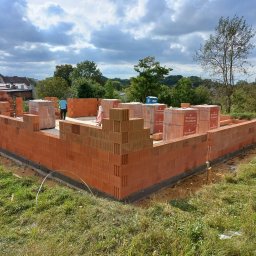 BARTZ Bartłomiej Wylężek - Wznoszenie ścian parteru domu z czerwonej cegły Porotherm, widoczny pracownik w niebieskiej koszulce i palety z cegłami, w tle zieleń i zabudowania mieszkalne pod pochmurnym niebem.