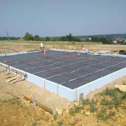 BARTZ Bartłomiej Wylężek - Fundament domu murowanego w trakcie budowy: styropianowe szalunki, zbrojenie z prętów stalowych i wystające rury kanalizacyjne na tle krajobrazu.