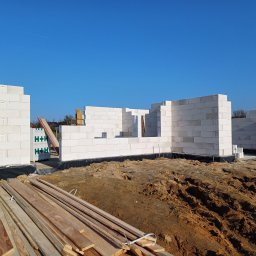 BARTZ Bartłomiej Wylężek - Wznoszenie ścian parterowego domu z białych bloczków na fundamencie z izolacją przeciwwilgociową, widoczne drewniane deski i palety materiałów budowlanych na placu budowy w słoneczny dzień.