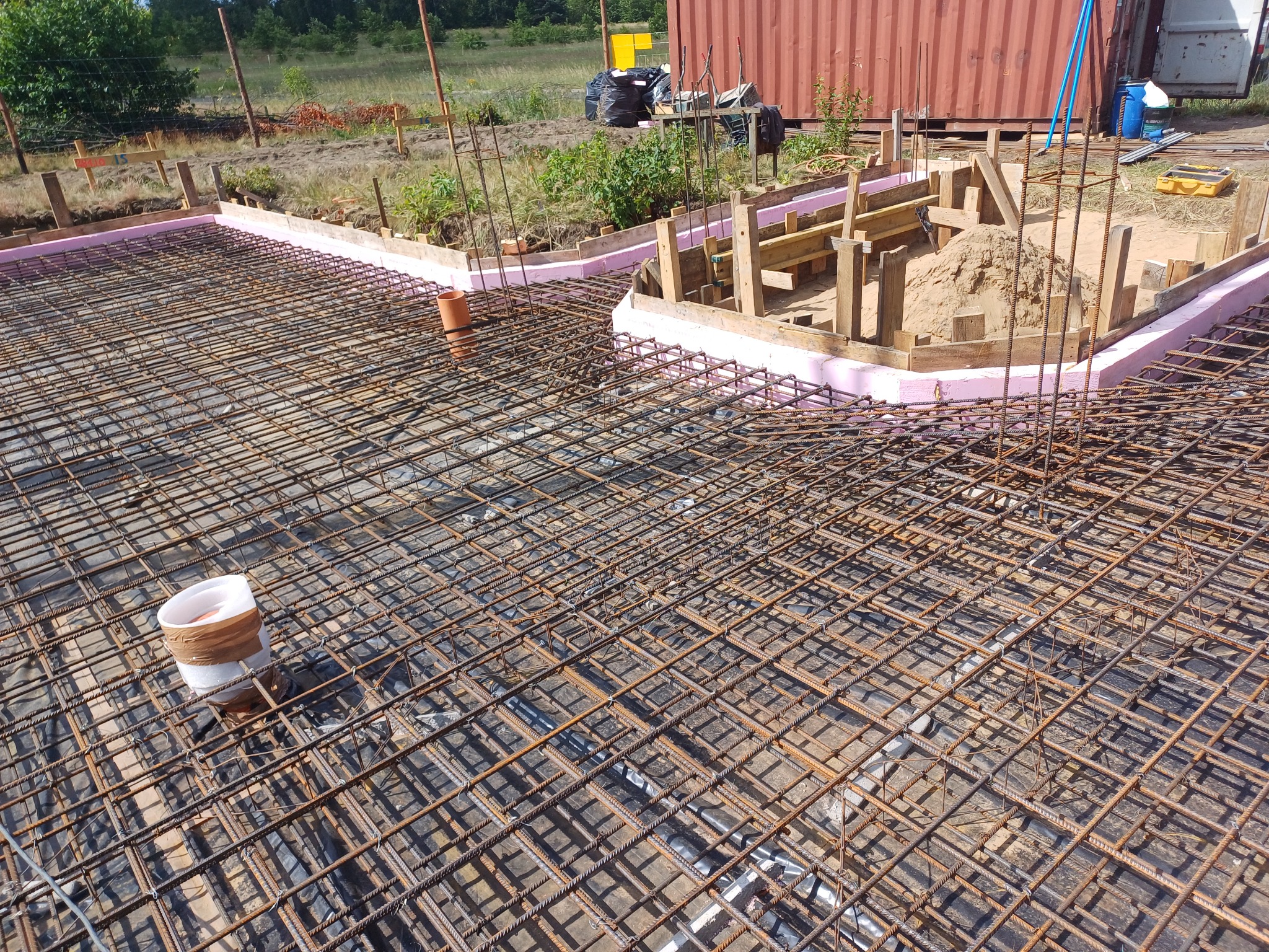 Przygotowanie zbrojenia pod płytę fundamentową domu jednorodzinnego z widoczną izolacją pionową i szalunkiem obwodowym, w tle widoczny kontener budowlany.