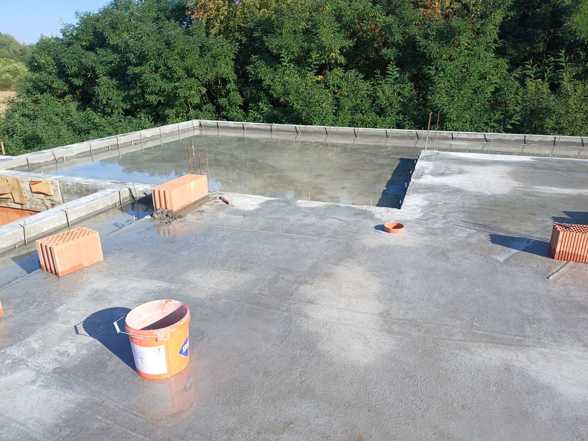 Wylany beton na stropie domu w trakcie budowy, widoczne bloczki ceramiczne, pręty zbrojeniowe oraz wiadro po zaprawie.