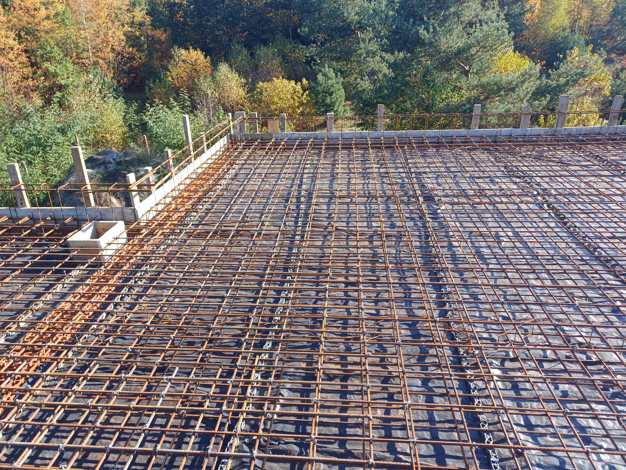Zbrojenie stropu ułożone na czarnej folii, widoczne drewniane szalunki i otaczający krajobraz leśny w jesiennej scenerii.