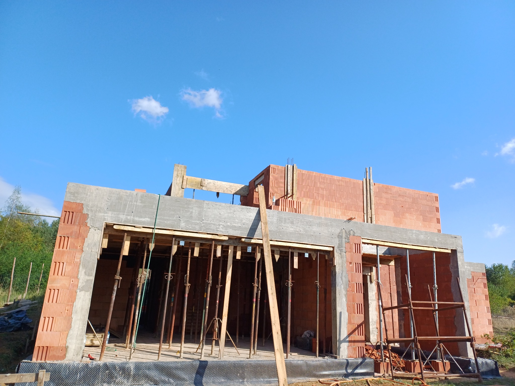 Budowa domu z czerwonej cegły ceramicznej Porotherm, widoczne szalunki i podpory stropu, niebieskie niebo w tle.