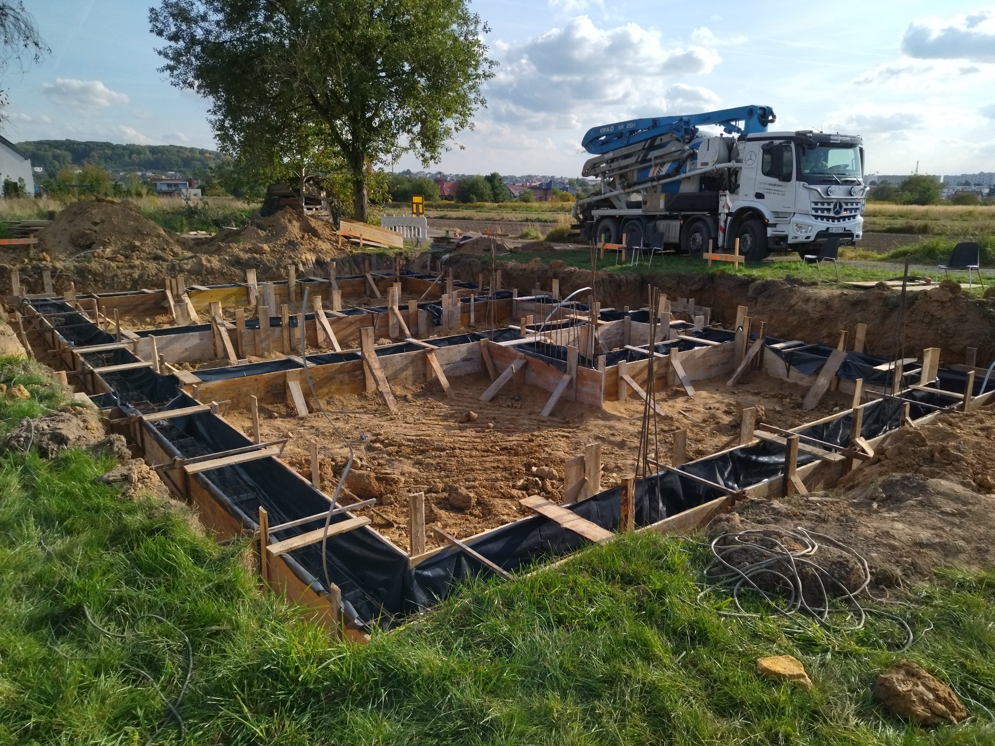 Przygotowanie fundamentów pod budowę domu: drewniane szalunki wyłożone folią, wystające pręty zbrojeniowe, w tle betonowóz z pompą do betonu i krajobraz wiejski.