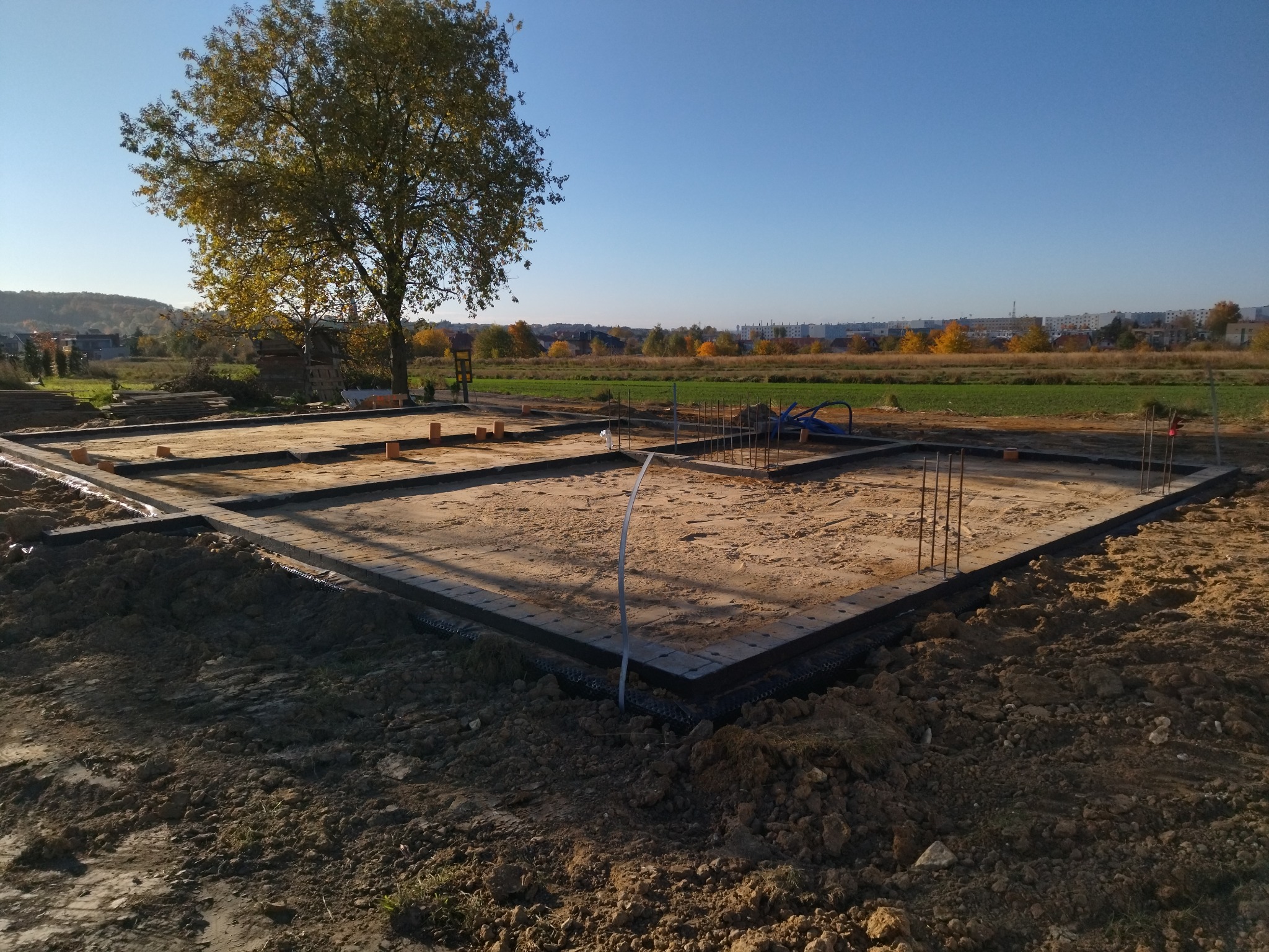 Fundamenty domu jednorodzinnego w trakcie budowy, widoczne zbrojenia i szalunki, w tle drzewo i zabudowa miejska pod błękitnym niebem.