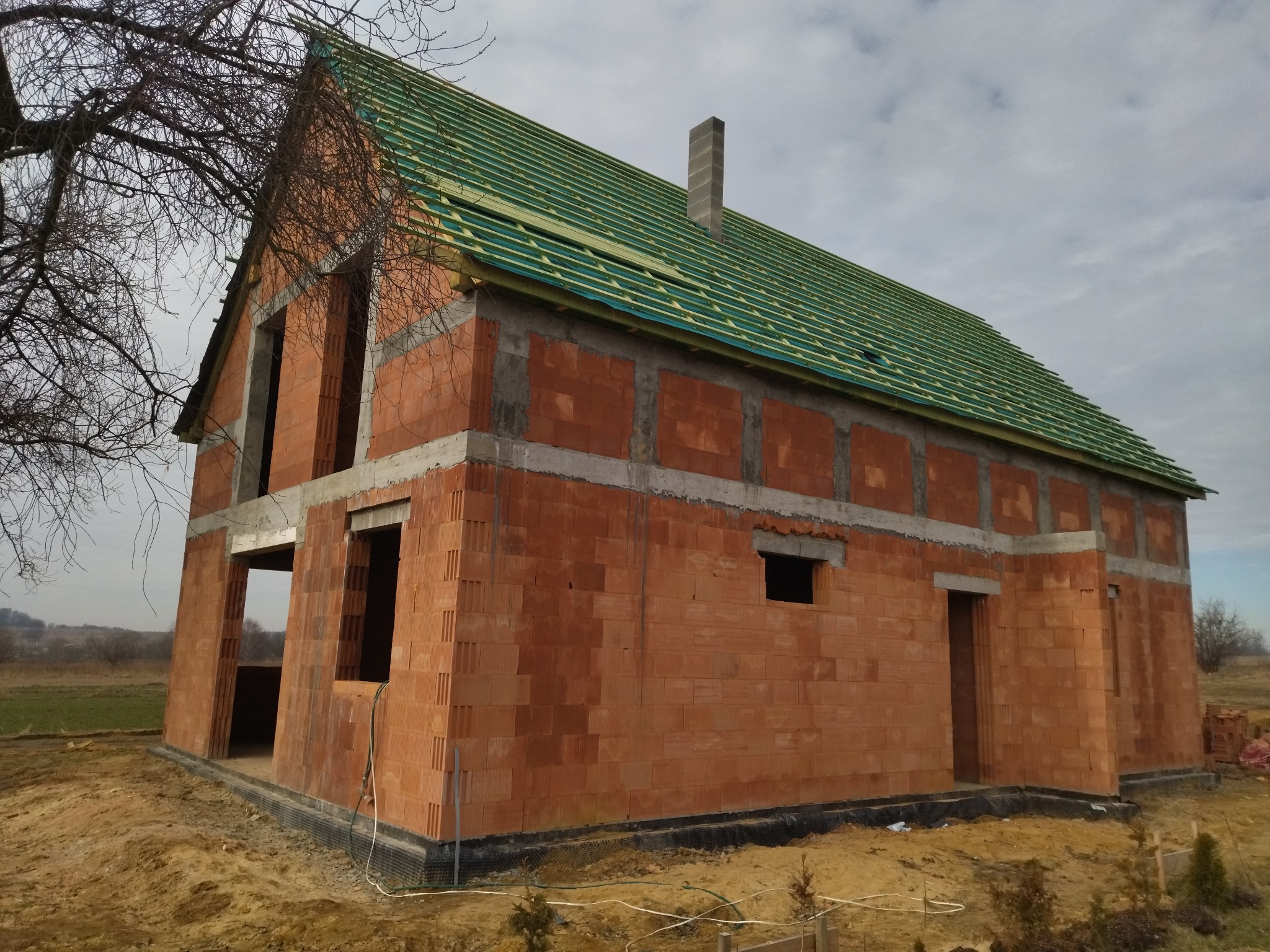 Surowy dom murowany z czerwonej cegły w trakcie budowy, z zielonym dachem i widocznym kominem, na tle pola i pochmurnego nieba.