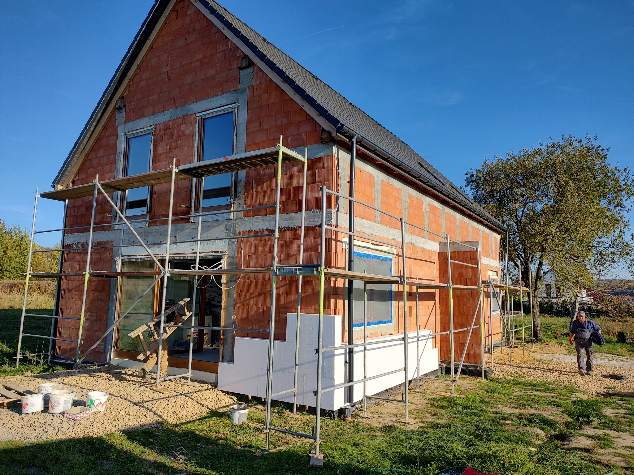 Surowy budynek z czerwonej cegły w trakcie ocieplania, z widocznym rusztowaniem i częściowo zamontowanym białym styropianem na elewacji, pracownik w oddali.