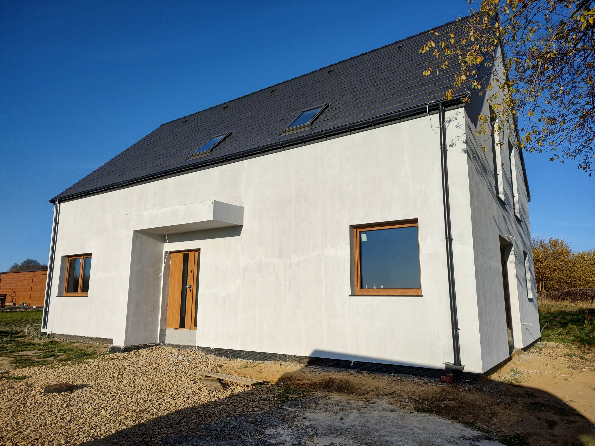 Nowoczesny dom jednorodzinny z białym tynkiem i ciemną dachówką, widoczne okna dachowe i drewniana stolarka okienna, teren wokół domu częściowo zagospodarowany kamieniami.