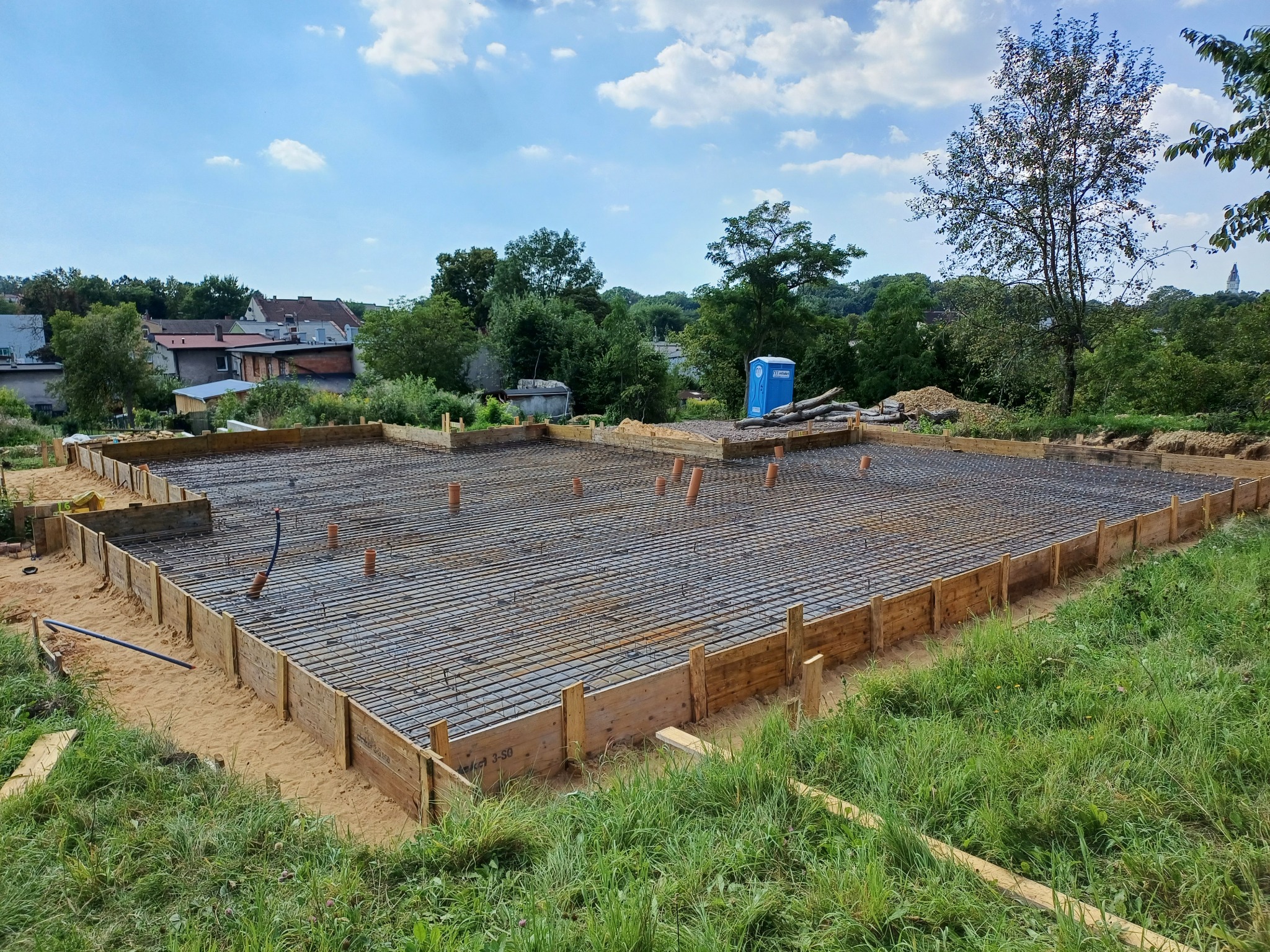 Przygotowanie zbrojenia i szalunku pod płytę fundamentową domu jednorodzinnego, widoczne wystające rury kanalizacyjne, w tle zabudowa wiejska i błękitne niebo z chmurami.