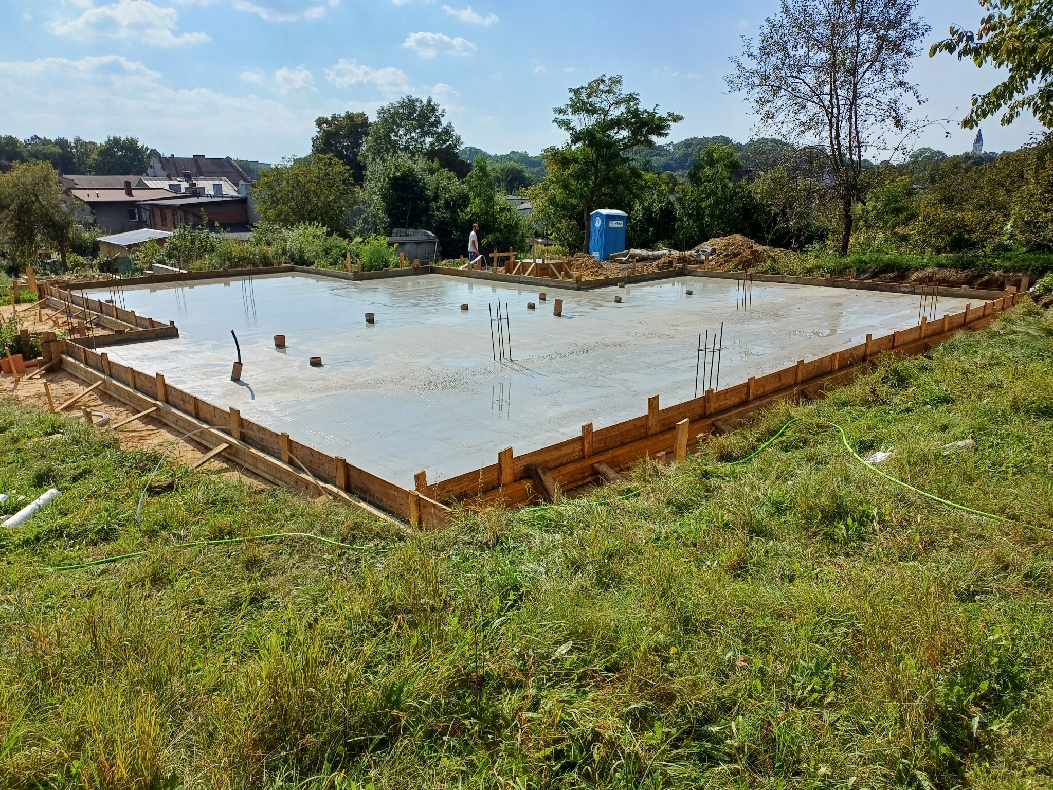 Wylana płyta fundamentowa pod budowę domu, otoczona drewnianym szalunkiem i z wystającymi prętami zbrojeniowymi, widok z góry na tle zieleni i zabudowań.