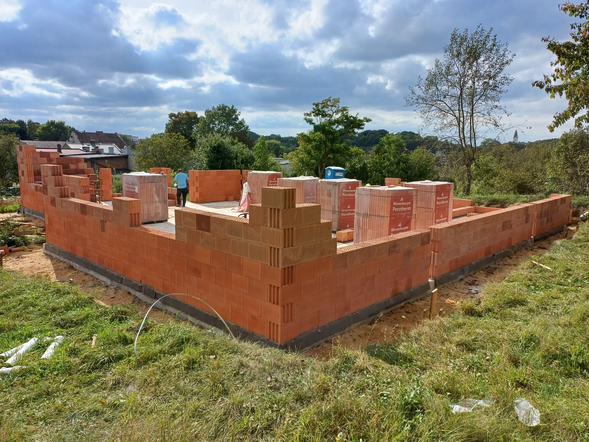 Wznoszenie ścian parteru domu z czerwonej cegły Porotherm, widoczny pracownik w niebieskiej koszulce i palety z cegłami, w tle zieleń i zabudowania mieszkalne pod pochmurnym niebem.