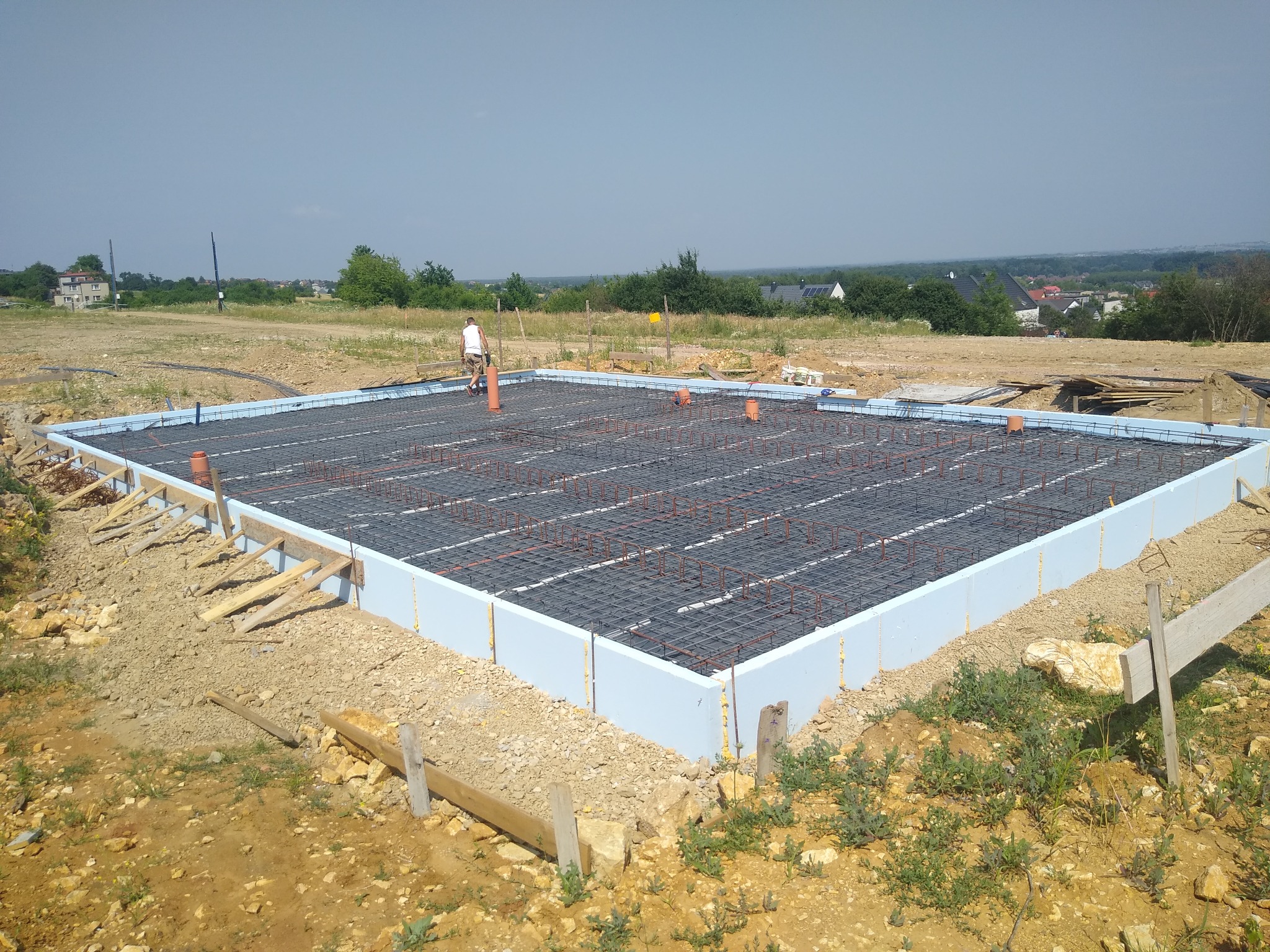 Fundament domu murowanego w trakcie budowy: styropianowe szalunki, zbrojenie z prętów stalowych i wystające rury kanalizacyjne na tle krajobrazu.