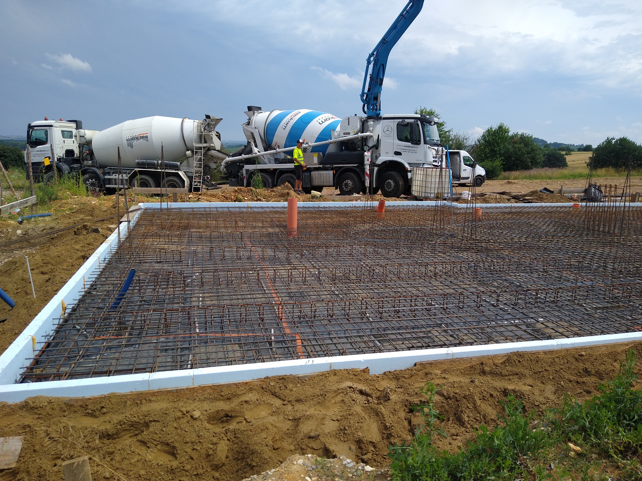 Zbrojenie fundamentu w szalunku ze styropianu, widoczne dwie betoniarki i pracownicy w tle.