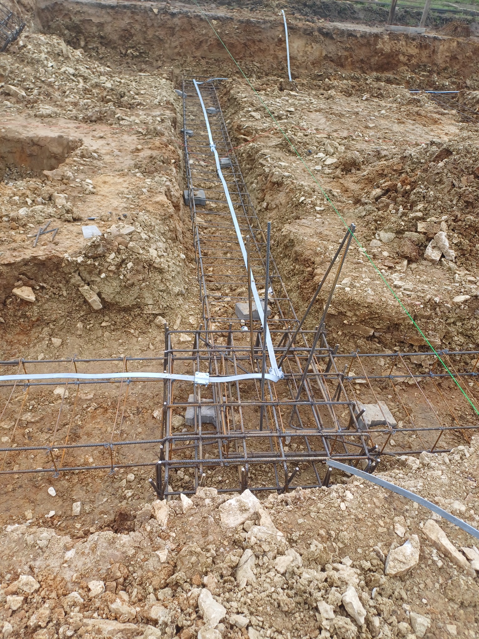 Zbrojenie fundamentów domu jednorodzinnego, widoczne stalowe pręty połączone w siatkę, częściowo zalane błotem, na tle wykopu ziemnego z kamieniami.
