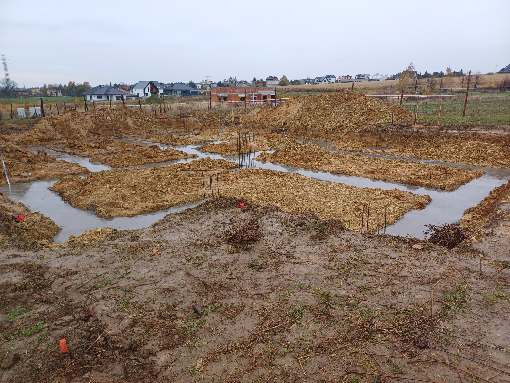 Wylane fundamenty domu jednorodzinnego z wystającymi prętami zbrojeniowymi, otoczone wykopaną ziemią i widokiem na zabudowę mieszkalną w tle w pochmurny dzień.