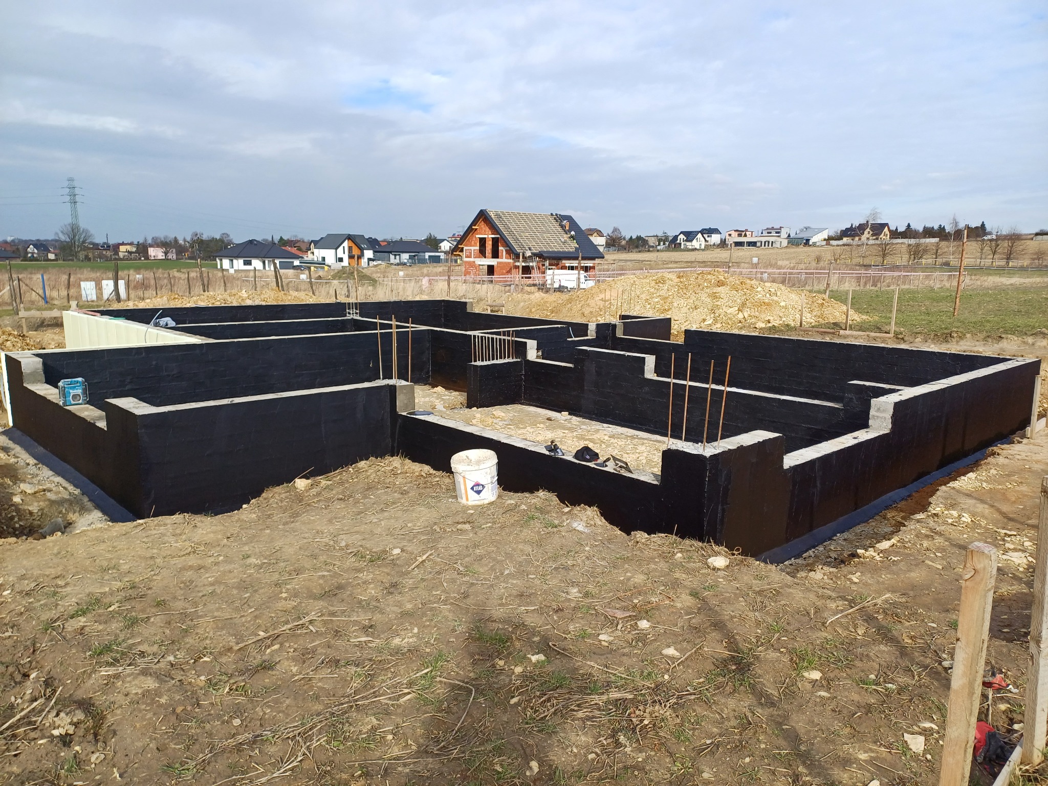 Fundamenty domu jednorodzinnego zabezpieczone czarną izolacją bitumiczną, widoczny biały pojemnik po masie izolacyjnej, w tle budowa domu w stanie surowym otwartym oraz zabudowa jednorodzinna.