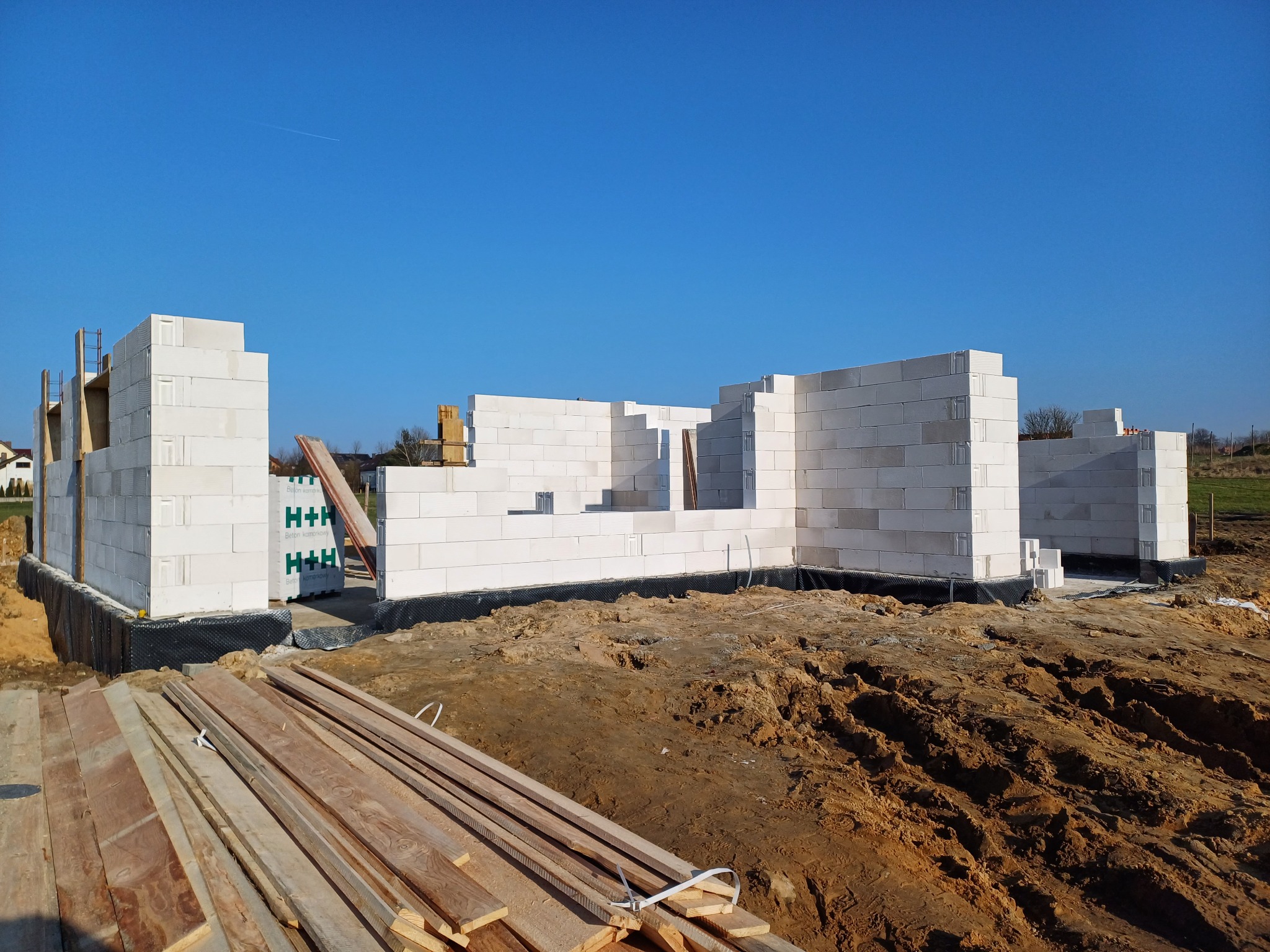 Wznoszenie ścian parterowego domu z białych bloczków na fundamencie z izolacją przeciwwilgociową, widoczne drewniane deski i palety materiałów budowlanych na placu budowy w słoneczny dzień.