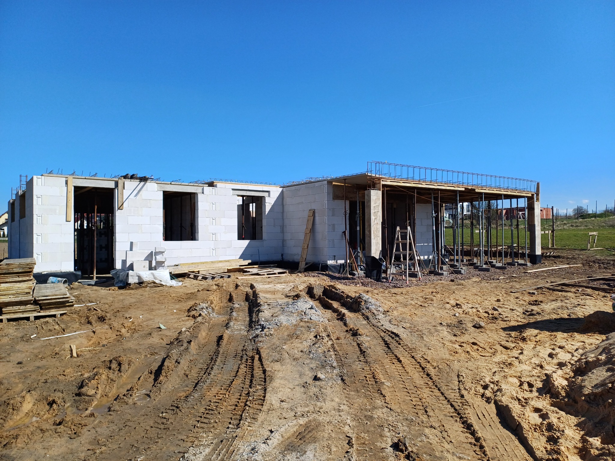 Budowa domu jednorodzinnego z białych bloczków, widoczne ślady opon na placu budowy i elementy konstrukcji stropu.