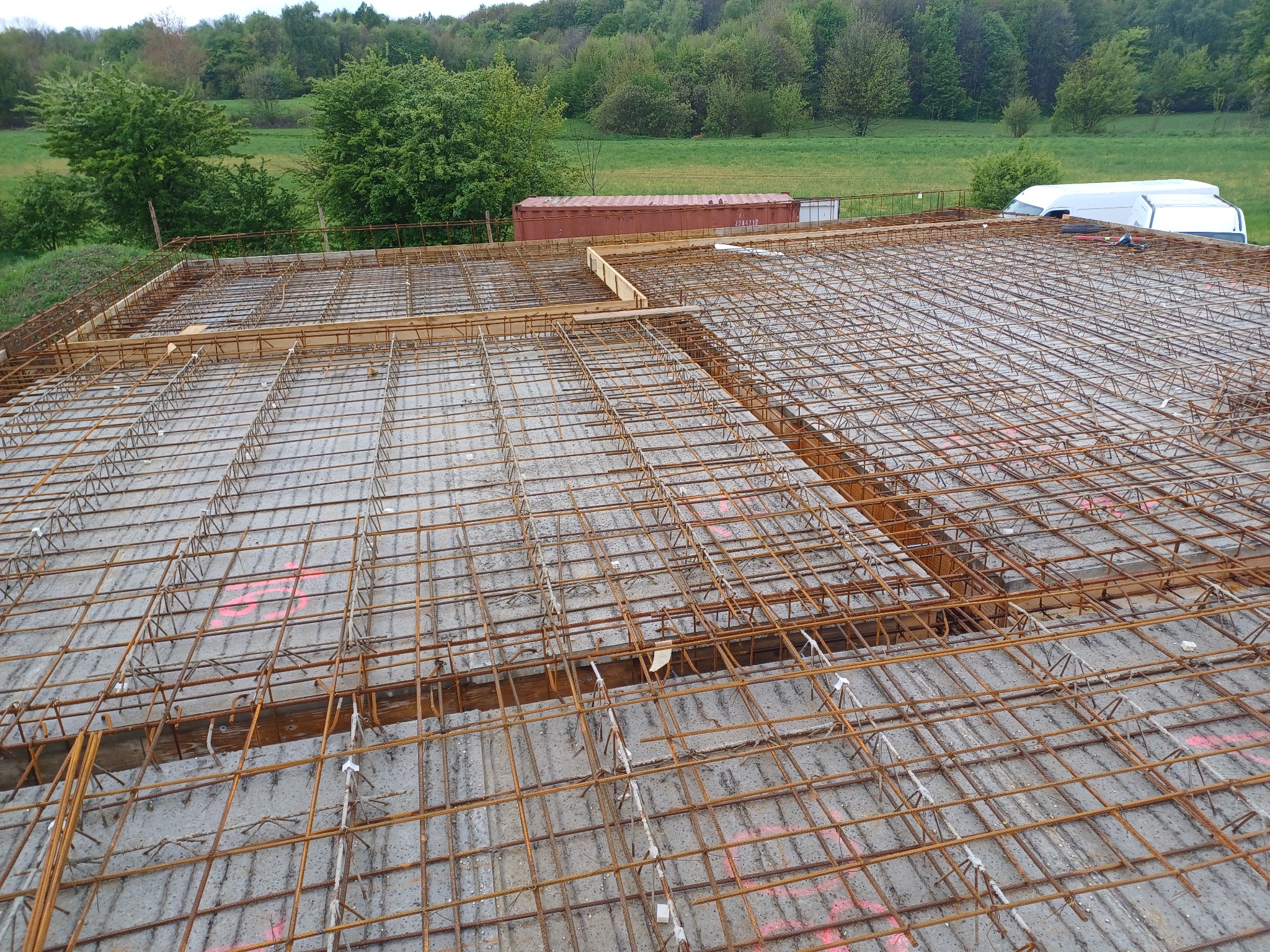 Zbrojenie płyty fundamentowej z widocznymi drewnianymi szalunkami i betonem, na tle zielonych drzew i nieba.
