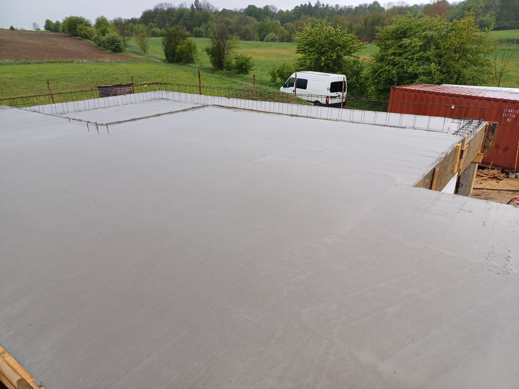 Świeżo wylana, gładka płyta betonowa z wystającymi prętami zbrojeniowymi, widok z góry na tle zieleni, białego busa i czerwonego kontenera.