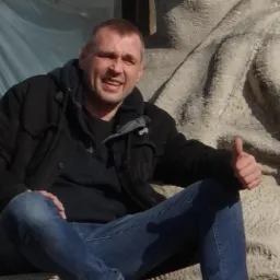 Mężczyzna w czarnej kurtce siedzi na betonowej konstrukcji, pokazując kciuk w górę.