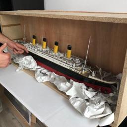 SIXPAK.PL Przeprowadzki - Model statku Titanic zabezpieczony w drewnianej skrzyni, przygotowany do transportu, z widoczną dłonią osoby dającej znak kciukiem w górę.