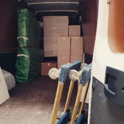 Wnętrze furgonetki załadowane kartonami, meblami owiniętymi folią i w&oacute;zkami transportowymi z niebieską wyści&oacute;łką, przygotowane do transportu.