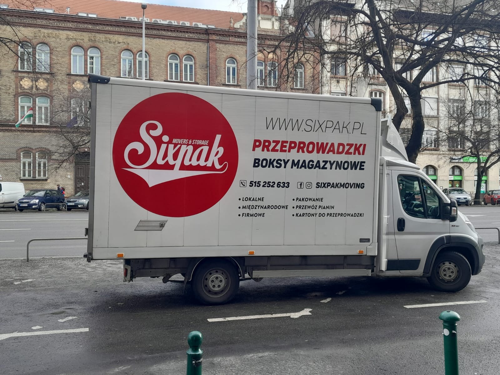 Samochód dostawczy firmy Sixpak Movers & Storage z Gdańska, z dużym czerwonym logo i informacjami kontaktowymi, zaparkowany na tle miejskiej zabudowy.