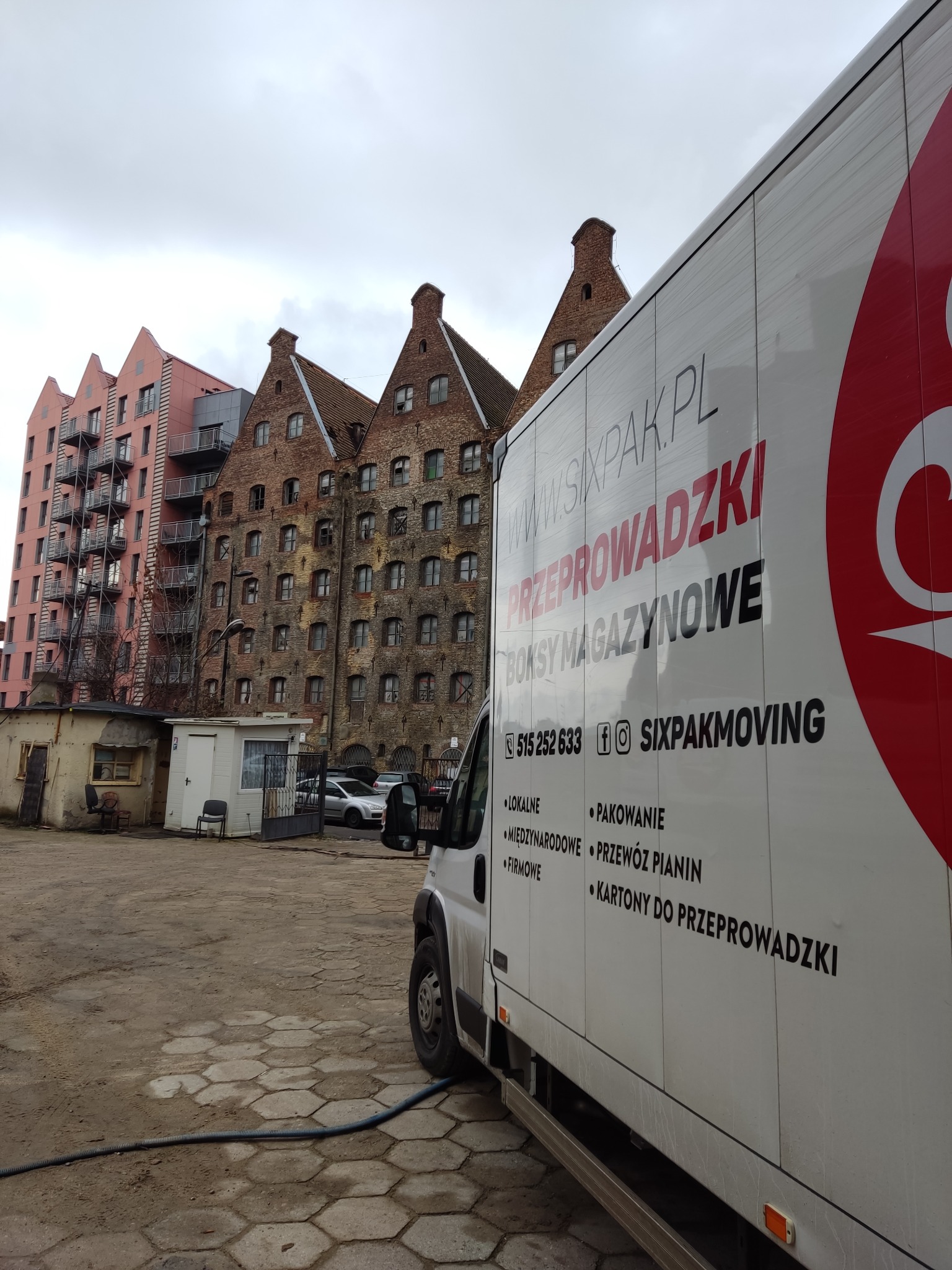 Samochód dostawczy firmy przeprowadzkowej z widocznym logo i danymi kontaktowymi na tle charakterystycznej architektury starego miasta.