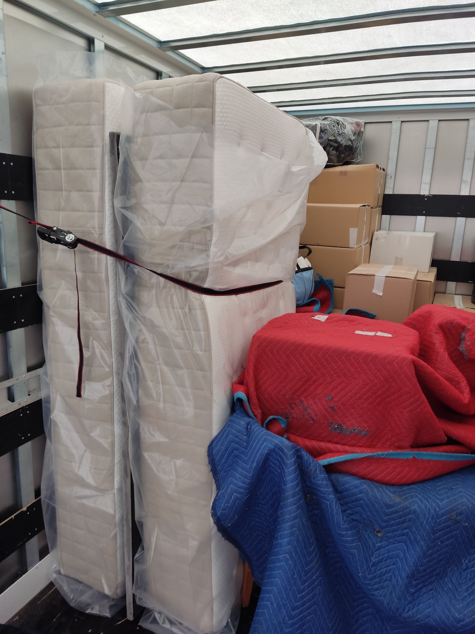 Wnętrze furgonetki załadowanej materacami owiniętymi folią, kartonami i meblami zabezpieczonymi czerwonymi i niebieskimi kocami transportowymi, spiętymi pasem mocującym.