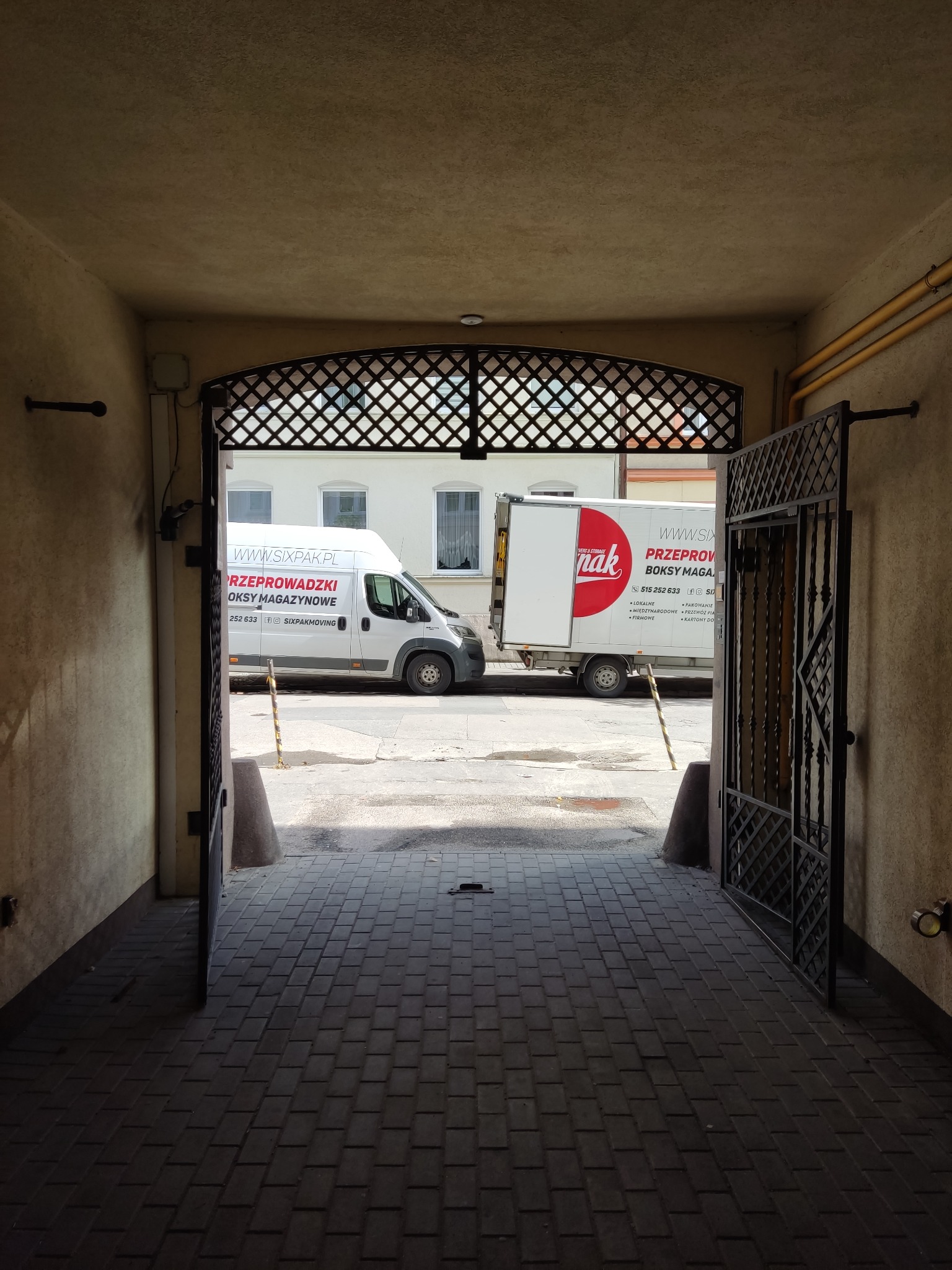 Dwa busy firmy przeprowadzkowej z widocznym logo i numerem telefonu, zaparkowane na zewnątrz, widziane przez otwartą bramę z metalową kratą w kamienicy.