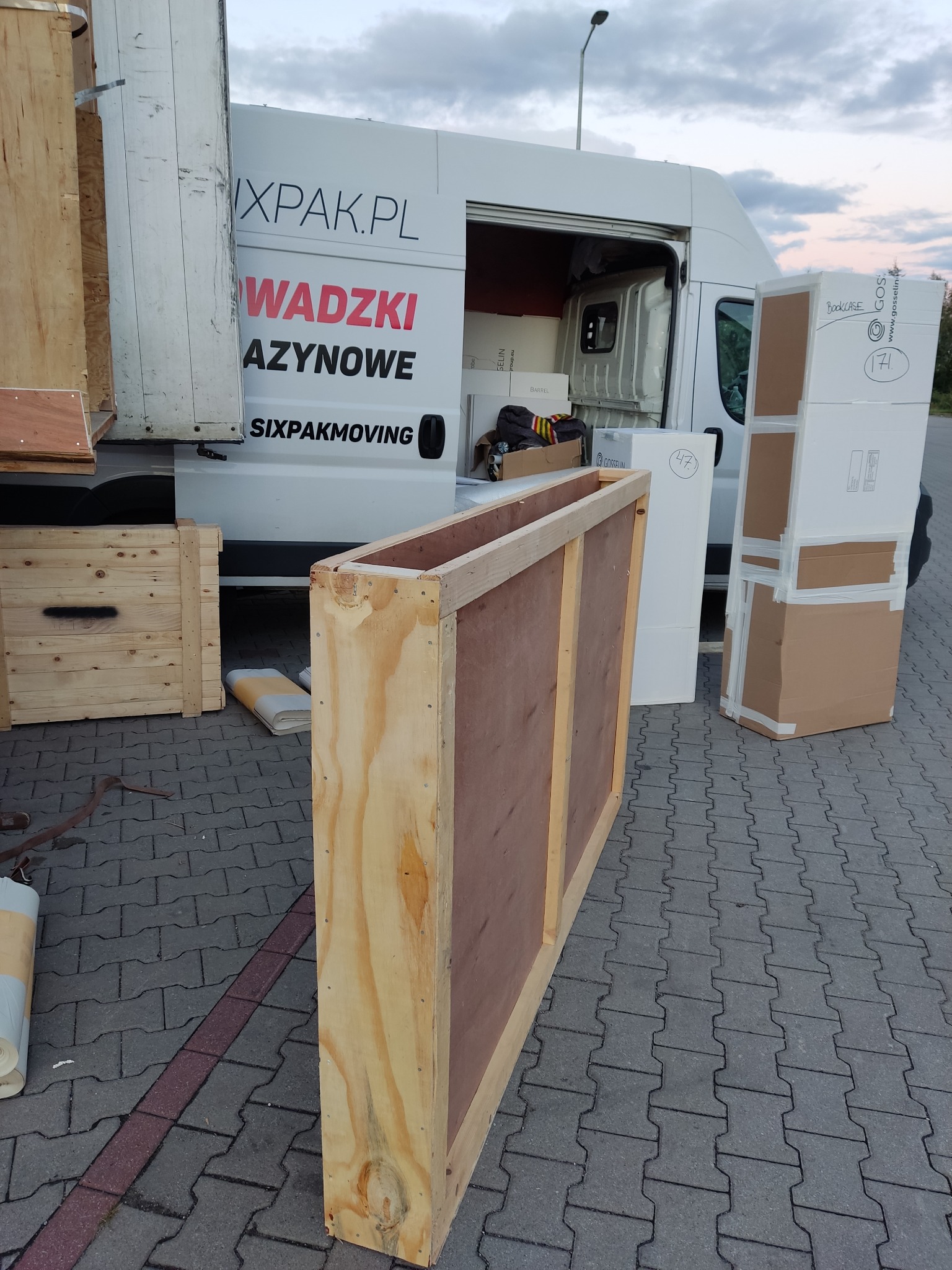 Otwarta skrzynia transportowa z jasnego drewna na tle częściowo załadowanego busa firmy SIXPAKMOVING, widoczne kartony i inne skrzynie gotowe do transportu.