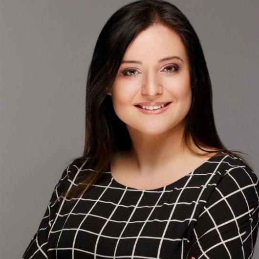 Portret uśmiechniętej kobiety z ciemnymi włosami, ubrana w czarno-białą sukienkę w kratę na szarym tle.