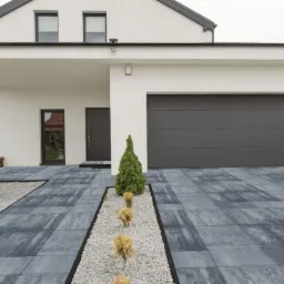 Nowoczesny dom z podjazdem wyłożonym szarą kostką brukową o nieregularnym wzorze, oddzielonym pasem drobnego żwiru i niskimi, ozdobnymi krzewami.