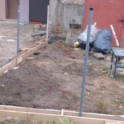 Piotr P - Przygotowanie terenu pod wylewkę fundamentów: drewniany szalunek, wykopana ziemia, pionowe rury, w tle czerwona brama garażowa i narzędzia.