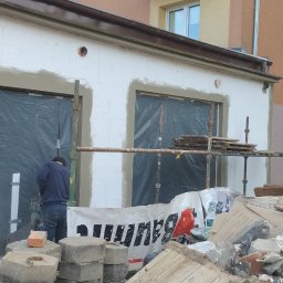 Piotr P - Pracownik w trakcie nakładania warstwy izolacyjnej na elewację budynku z przygotowanymi otworami okiennymi, widoczne rusztowania i gruz.