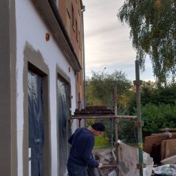 Piotr P - Pracownik w Charbrowie przygotowuje maszynę do natrysku tynku na elewacji budynku, widoczne rusztowanie i zabezpieczone okna folią.