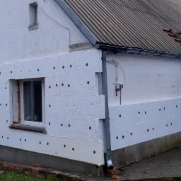 Fragment elewacji domu w trakcie nakładania izolacji termicznej, widoczne płyty styropianowe z czarnymi kołkami mocującymi, częściowo otynkowana ściana na górze, stara dachówka.
