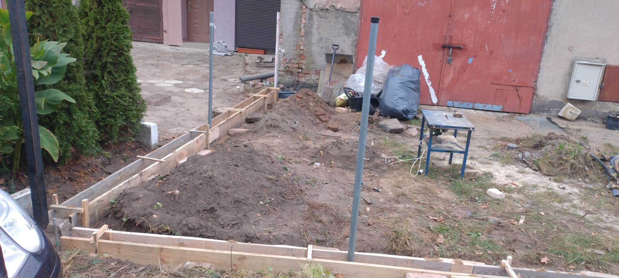 Przygotowanie terenu pod wylewkę fundamentów: drewniany szalunek, wykopana ziemia, pionowe rury, w tle czerwona brama garażowa i narzędzia.