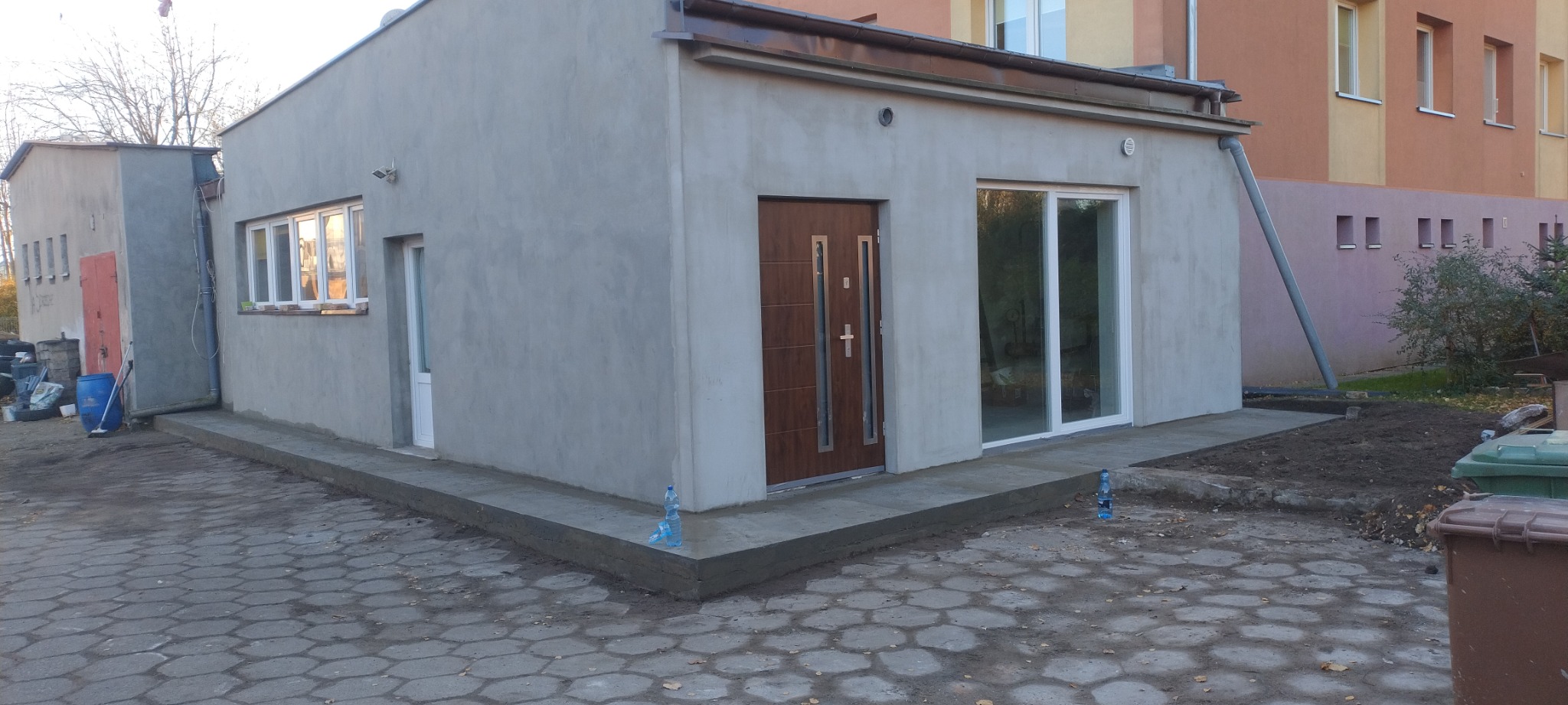 Budynek po szpachlowaniu i malowaniu elewacji zewnętrznej, widoczna nowa stolarka okienna i drzwiowa, teren wokół budynku częściowo zagospodarowany.