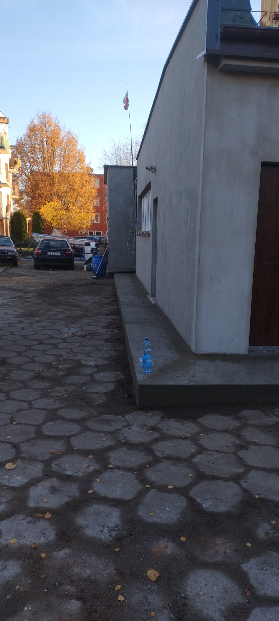 Budynek po nałożeniu szarego tynku zewnętrznego, z widocznymi butelkami wody na świeżo wykończonym cokole, w tle samochody i drzewa z jesiennymi liśćmi.