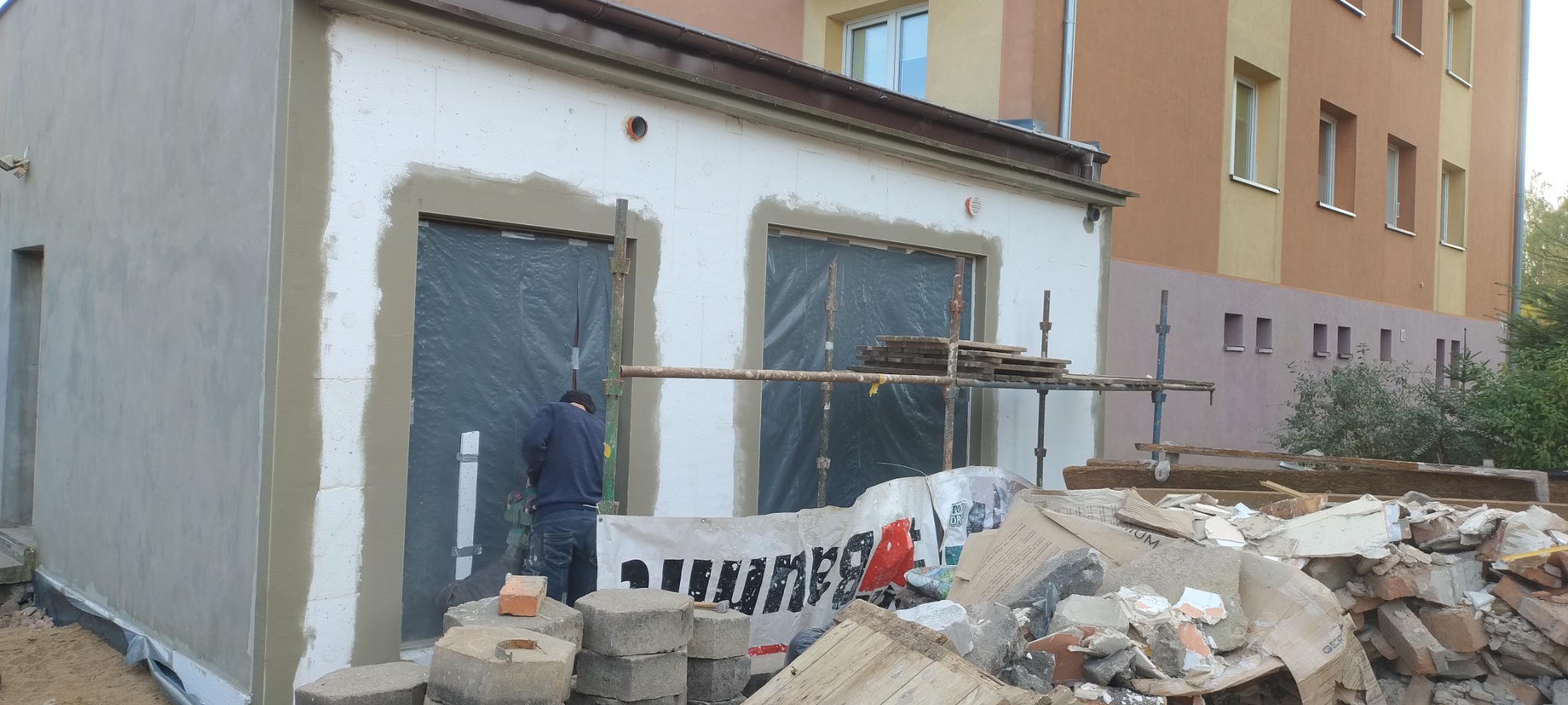 Pracownik w trakcie nakładania warstwy izolacyjnej na elewację budynku z przygotowanymi otworami okiennymi, widoczne rusztowania i gruz.