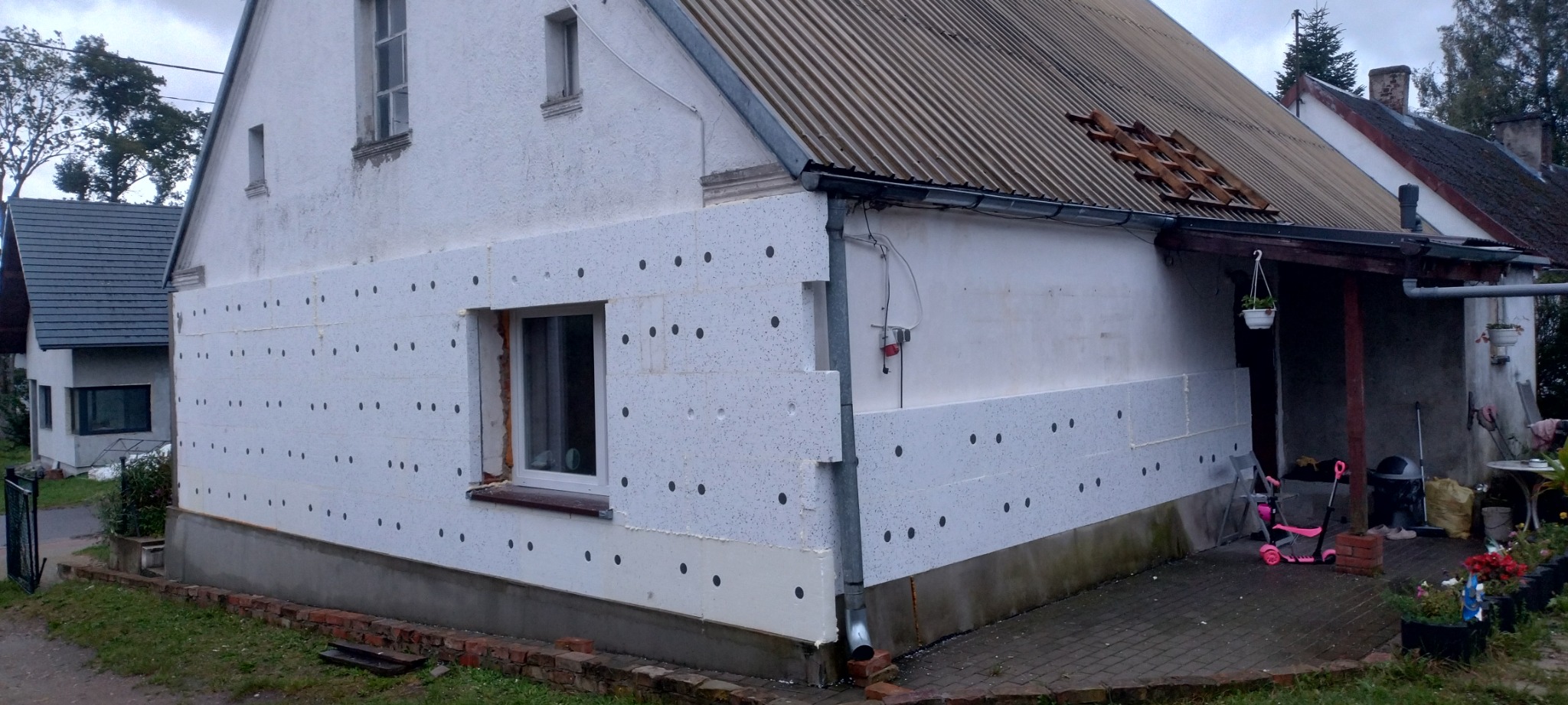 Fragment elewacji domu w trakcie nakładania izolacji termicznej, widoczne płyty styropianowe z czarnymi kołkami mocującymi, częściowo otynkowana ściana na górze, stara dachówka.