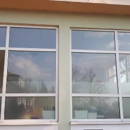 Dwa duże okna z białymi ramami, jedno z nich uchylone, odbijające błękitne niebo i drzewa.