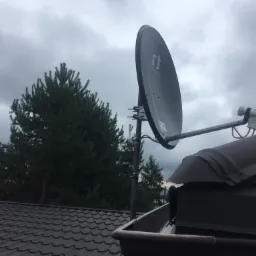 Szara antena satelitarna Inverto zamontowana na metalowym dachu z widocznym drzewem iglastym i pochmurnym niebem w tle.